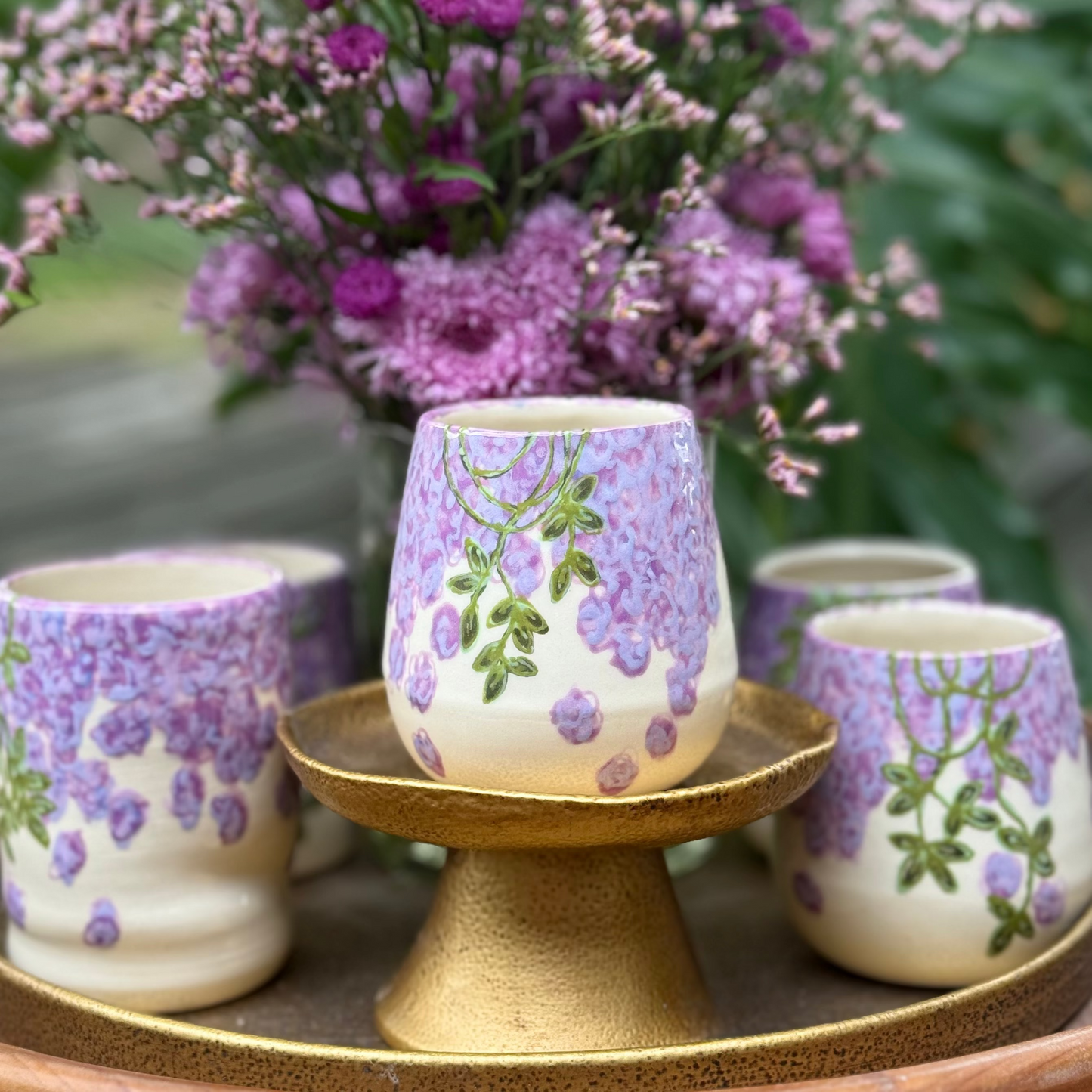 handmade and hand painted ceramic cups with handpainted wisteria, on a gold stand, surrounded by purple flowers of wisteria