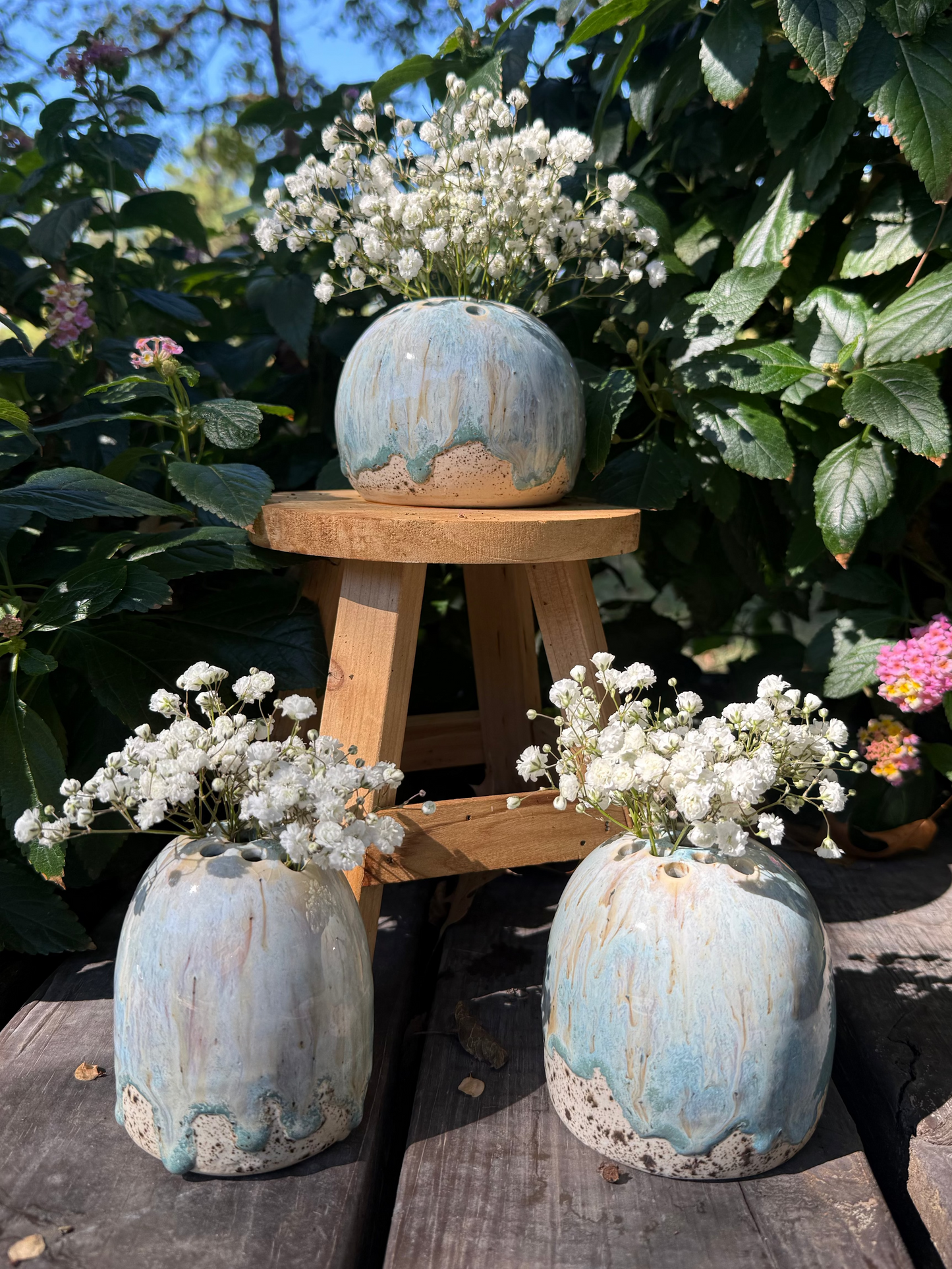 Three ceramic vases with a textured blue finish, each containing small white flowers, on a wooden surface with green foliage in the background.
