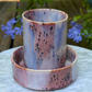 A handmade ceramic cup and saucer set with a marbled finish in shades of pink, purple, and blue, featuring dark speckled accents, displayed outdoors on a white marble surface with blurred purple flowers and green foliage in the background.