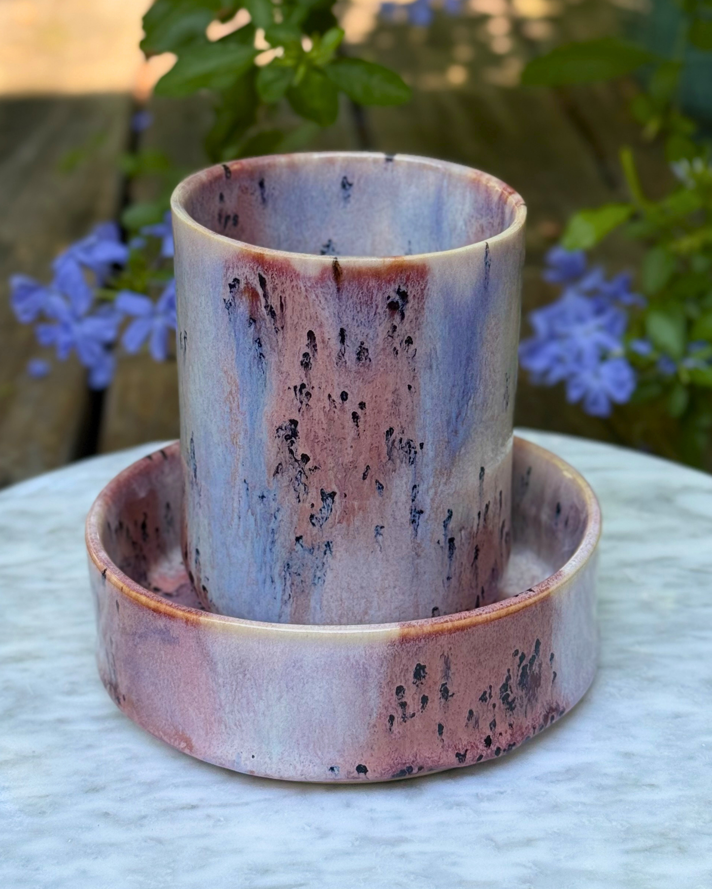 A handmade ceramic cup and saucer set with a marbled finish in shades of pink, purple, and blue, featuring dark speckled accents, displayed outdoors on a white marble surface with blurred purple flowers and green foliage in the background.