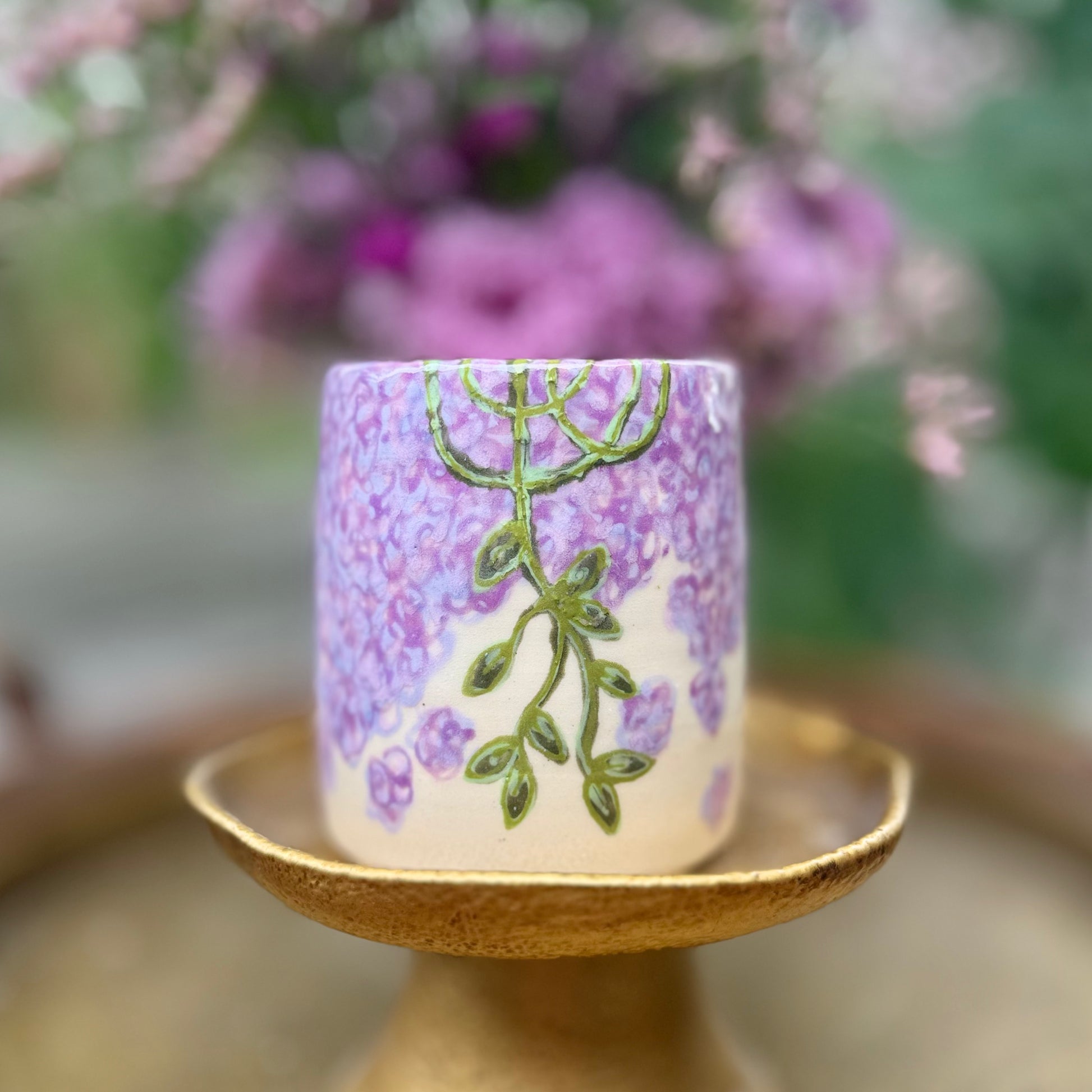 handmade and hand painted ceramic cup with handpainted wisteria, on a gold stand, surrounded by purple flowers of wisteria