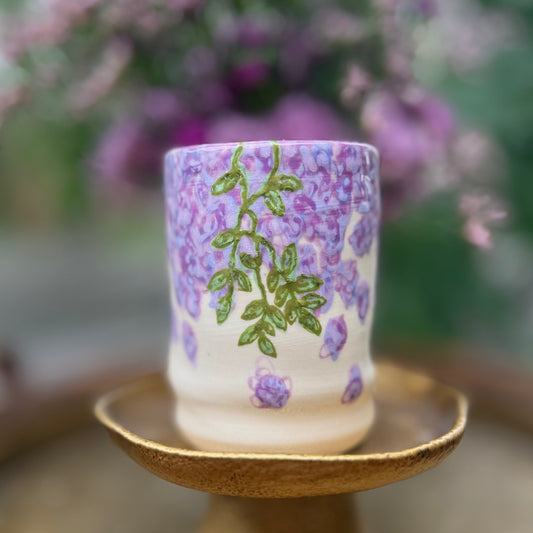 handmade and hand painted ceramic cup with handpainted wisteria, on a gold stand, surrounded by purple flowers of wisteria