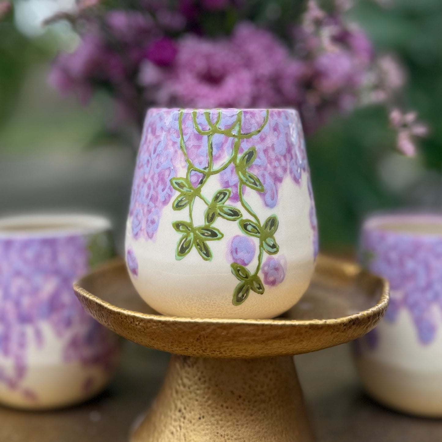 handmade and hand painted ceramic cups with handpainted wisteria, on a gold stand, surrounded by purple flowers of wisteria