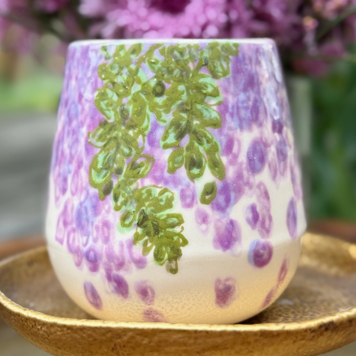 handmade and hand painted ceramic cup with handpainted wisteria, on a gold stand, surrounded by purple flowers of wisteria