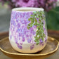 handmade and hand painted ceramic cup with handpainted wisteria, on a gold stand, surrounded by purple flowers of wisteria