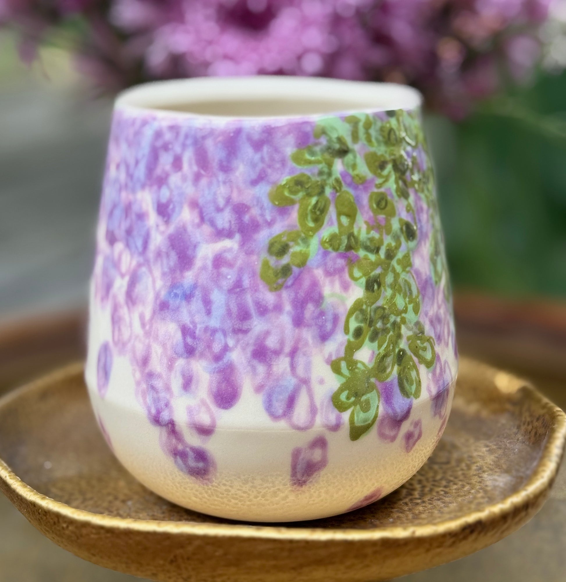 handmade and hand painted ceramic cup with handpainted wisteria, on a gold stand, surrounded by purple flowers of wisteria