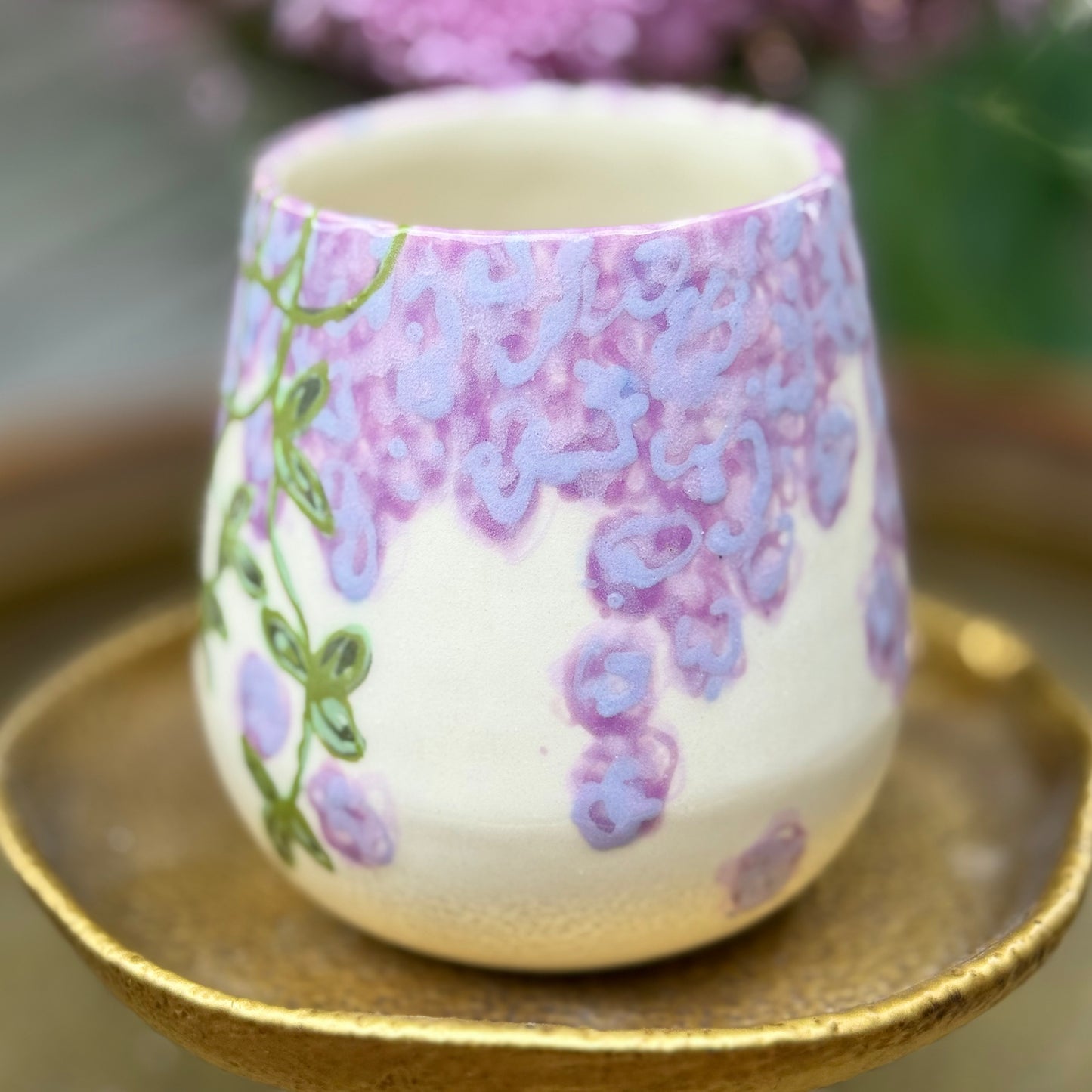handmade and hand painted ceramic cup with handpainted wisteria, on a gold stand, surrounded by purple flowers of wisteria