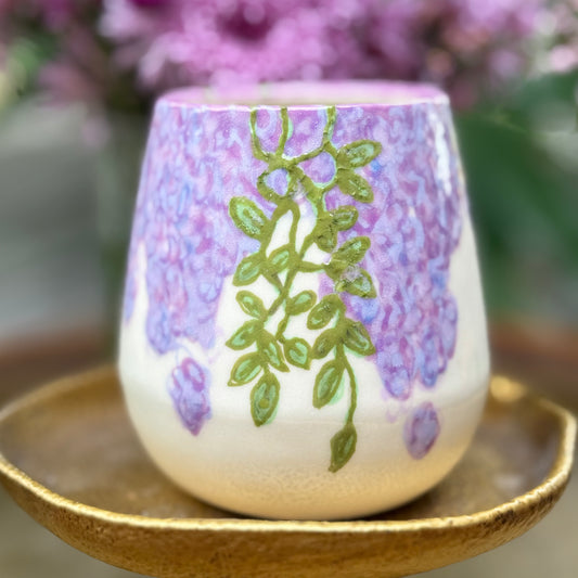handmade and hand painted ceramic cup with handpainted wisteria, on a gold stand, surrounded by purple flowers of wisteria
