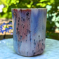 A handmade ceramic cup with a marbled pattern of blue, pink, and gray, featuring small black speckles, set on a light marble surface with a blurred green garden background.