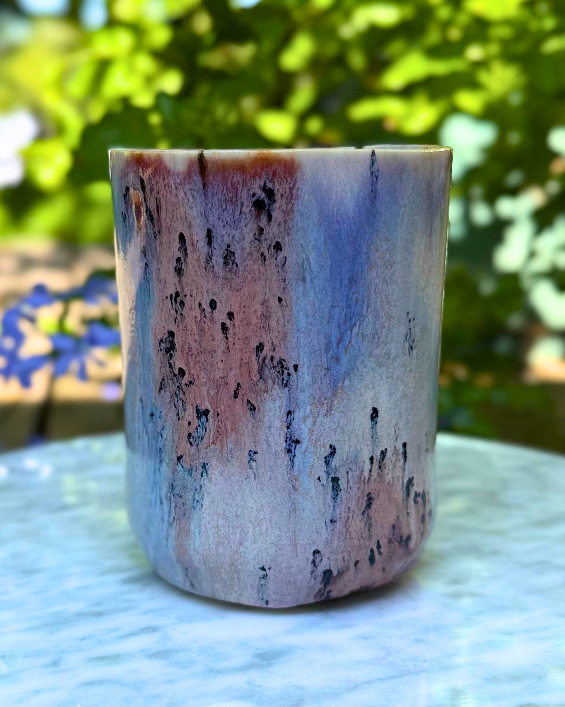 A handmade ceramic cup with a marbled pattern of blue, pink, and gray, featuring small black speckles, set on a light marble surface with a blurred green garden background.